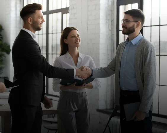 Three people in an office. Two men, one in a dark suit, another in a casual sweater, shake hands, smiling.