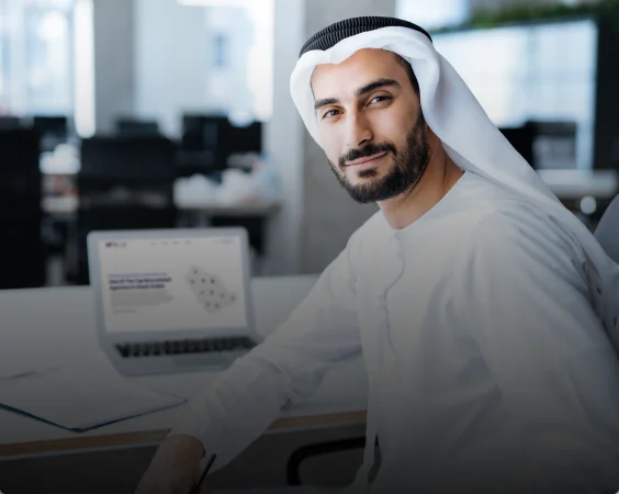 A man in traditional Middle Eastern attire, including a white kandura and gutra, sits at a desk with an open laptop in front of him in a modern office setting.