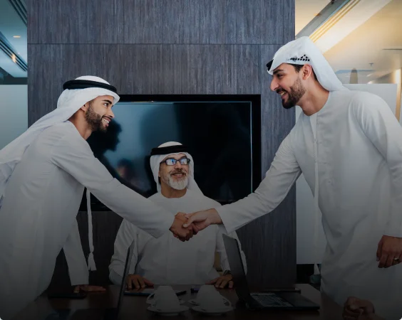 Three men dressed in traditional Middle Eastern attire are in an office setting. Two men are shaking hands and smiling, while the third man, seated at a table behind them, watches their interaction with a content expression.