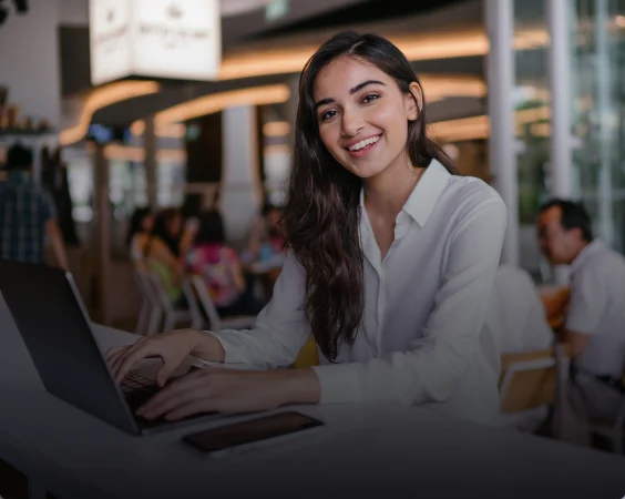 A young woman with long dark hair, wearing a white blouse, smiles while working on a laptop in a modern, brightly lit cafe.