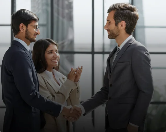 Two men in business suits shake hands while smiling at each other, with a woman standing between them, clapping and also smiling. The atmosphere suggests a successful partnership with an HR outsourcing and recruitment agency from Pakistan.