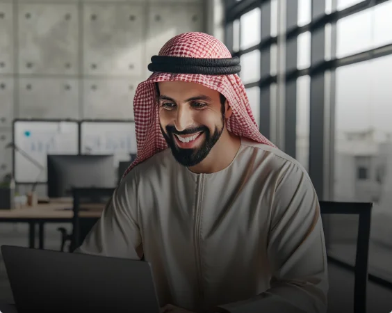 A smiling man in a traditional Middle Eastern attire, including a red and white checkered keffiyeh, works on a laptop in a modern office with large windows and charts depicting business growth on the wall in the background.