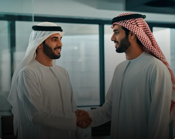 Two men in traditional Middle Eastern attire are shaking hands and smiling in an office setting with glass partitions.