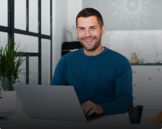 A man with a beard smiles while using a laptop in a modern, brightly lit office. He is wearing a blue sweater and sitting at a desk with a small black coffee cup and a potted plant.