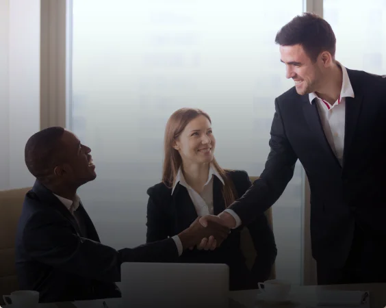 Three people in business attire are interacting in an office setting. One man is seated and shaking hands with another man who is standing; a woman, involved in HR outsourcing.